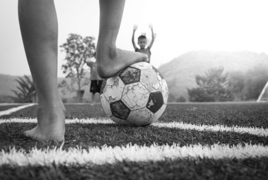 Boy with foot on well used soccer ball. Boy in background holds up arms. Black and white photo.