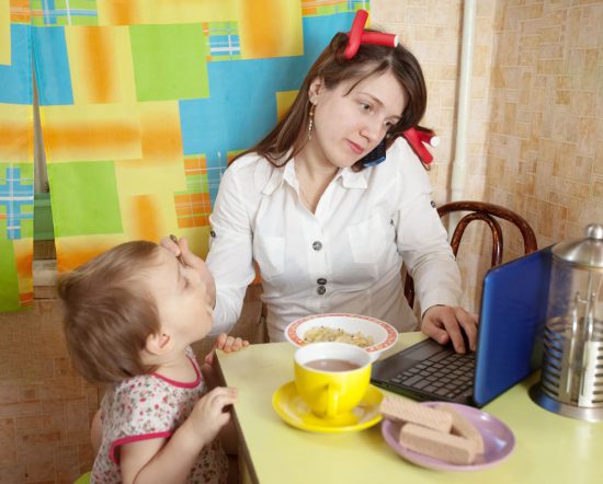 Mother feeds child while looking at her computer screen, distracted parenting