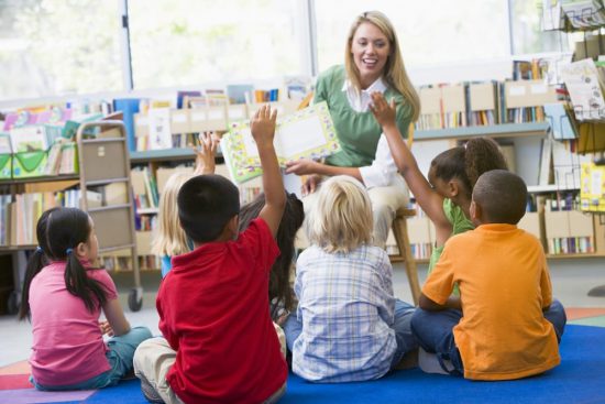 Smiling blonde teacher with either young students raising hand, from behind National Teacher Appreciation Day