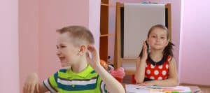 Squirmy Boy with ADHD seated at desk in front of girl politely raising her hand shows it's hard to manage ADHD in the classroom