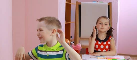 Squirmy Boy with ADHD seated at desk in front of girl politely raising her hand shows it's hard to manage ADHD in the classroom