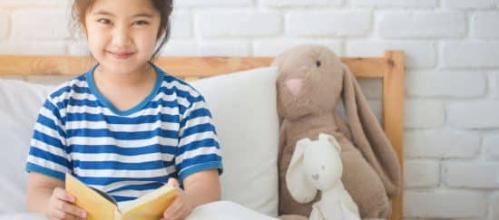 Sleep Duration a Factor in Childhood Obesity. Smiling Chinese Girl reading in bed, stuffed bunny nearby.