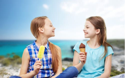 Two young girlfriends eating treats on the beach, smiling at each other.