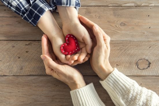 A woman's hands encircle a child's hands holding handmade red cloth heart