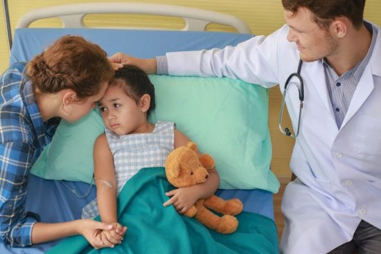 Mother holds hand of sick child in hospital bed as ER doctor in white coat with stethoscope looks on.