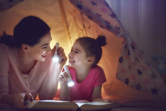 Mother Reads to Daughter in tent with both holding flashlights and smiling