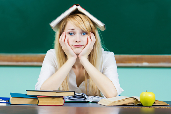 student with textbook upside down on head, chin in hands, illustrates the frustration experienced by some students in our age-based graduation system