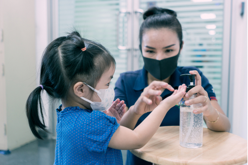 Masked mom helps masked daughter use hand sanitizer against the spike in child cases of COVID19