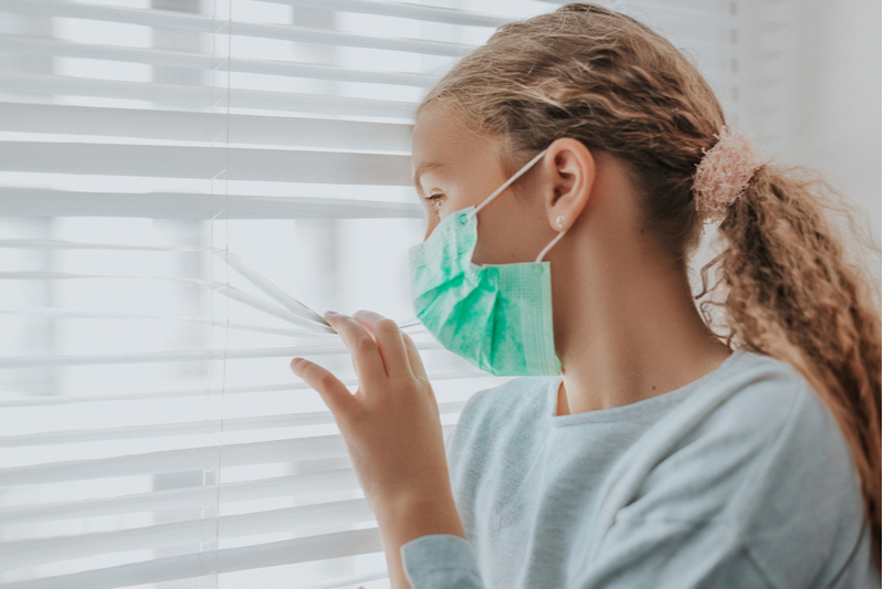 girl with Covid19 wearing mask peering out of window from bedroom quarantine during spike in child coronavirus cases