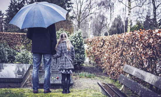 Little girl holds father hand at grave in rain under umbrella