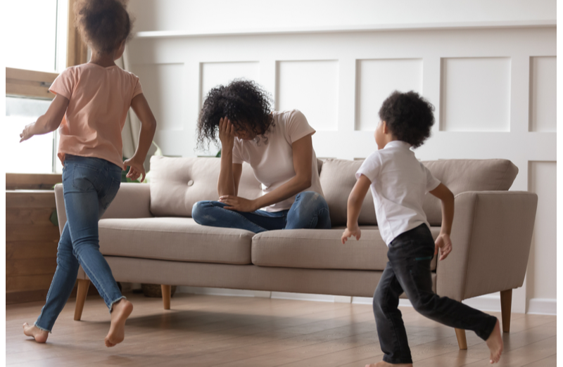 Children running around sofa with tired mom head in hands