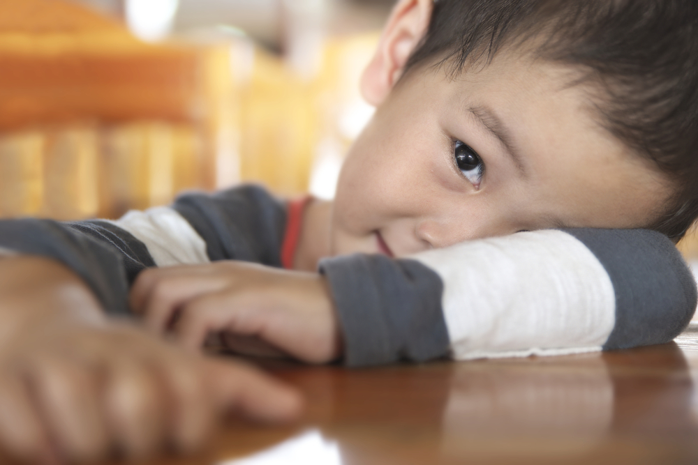 bashful asian boy hides eye with arm on table, peeks out