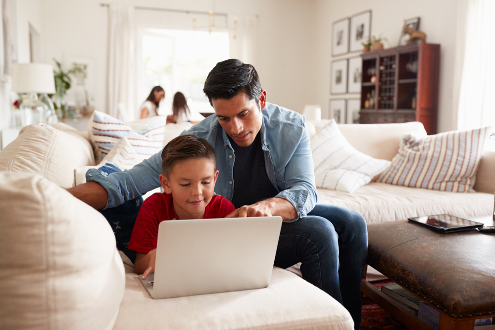 preteen boy lies on sofa father points something out on laptop screen, teaches about elections