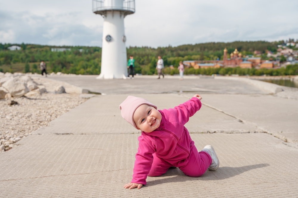 smiling baby bundled up in pink crawls, lighthouse in background