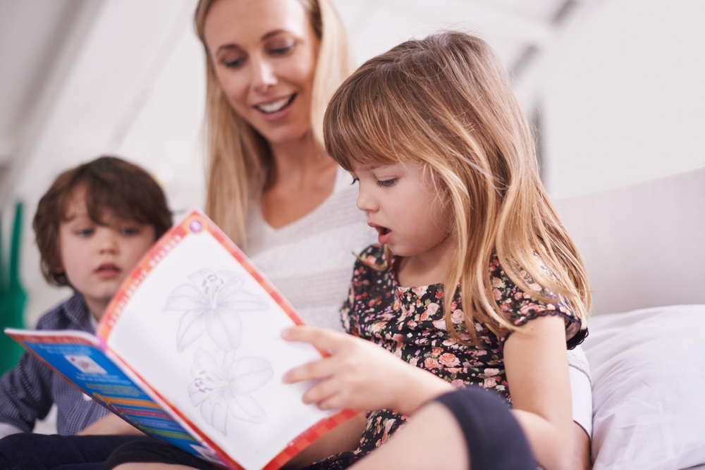 Five-year-old blond girl reads book to smiling mother with little brother looking on (How to Teach Children to Read at Any Age)