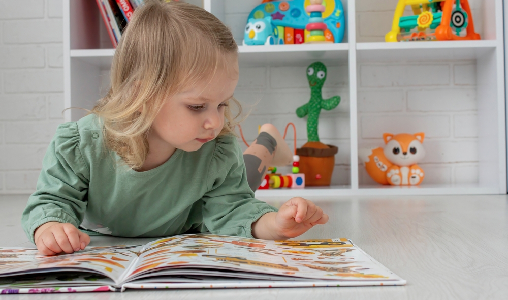 Toddler girl lying on the floor reading a picture book, with toys and books on a shelf behind her