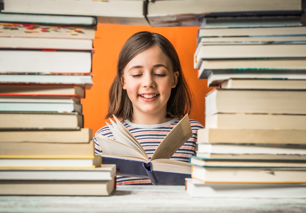 Smiling girl reading a book, framed by tall stacks of books on either side