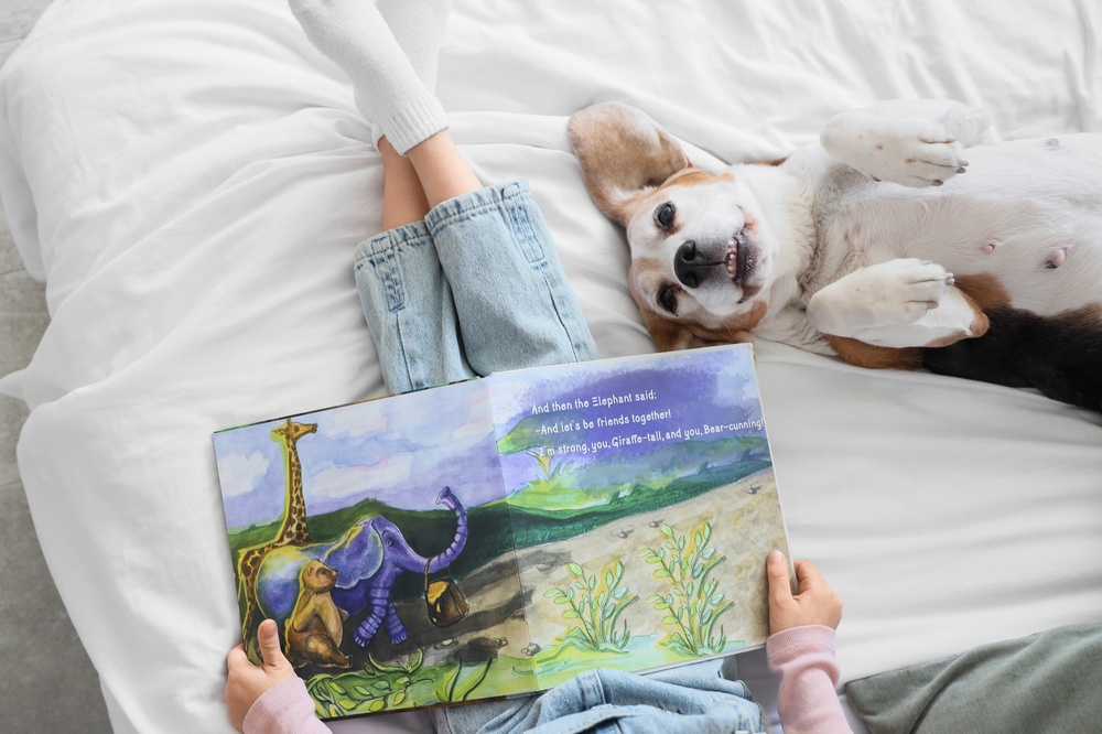 Overhead view of a child reading a picture book on a bed next to a beagle lying on its back